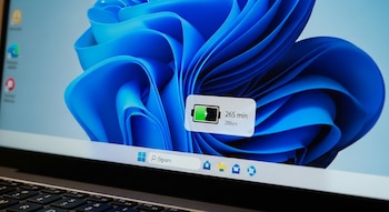 Una pantalla de computador portátil con Windows 11 que muestra un icono de batería verde con un rayo y el texto '265 min', sobre un fondo azul degradado.