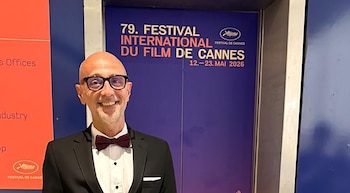 Hombre calvo con gafas y pajarita roja sonriendo, de pie frente a un cartel azul del 79. Festival Internacional de Cine de Cannes y un panel rojo a la izquierda