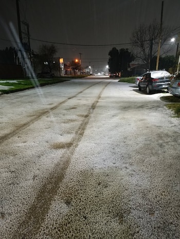 Calle asfaltada cubierta de granizo blanco de noche, con marcas de neumáticos en el centro y automóviles estacionados a la derecha