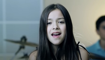 Primer plano de una niña de cabello largo y oscuro que canta, con una batería desenfocada y otra persona en el fondo