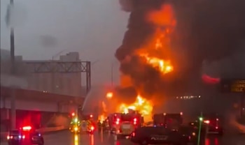 Imagen de una explosión y gran incendio con llamas anaranjadas y humo denso en una autopista, con camiones de bomberos y vehículos de policía con luces intermitentes