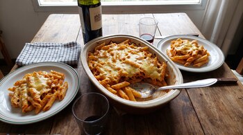 Fuente de cerámica con pasta penne gratinada, dos platos servidos, una botella de vino y dos vasos sobre una mesa de madera.
