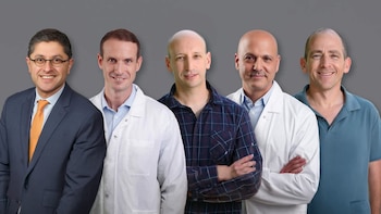 Cinco hombres sonriendo de pie frente a un fondo gris. Usan traje, batas de laboratorio, camisa a cuadros y polo
