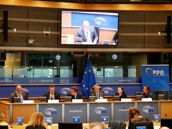 Salón de conferencias con un panel de seis personas en una mesa principal, audiencia sentada, bandera de la Unión Europea y una pantalla LED grande