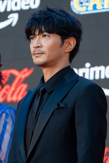 Kenjiro Tsuda, vestido con traje, camisa y corbata negras, posa ante un fondo oscuro con anuncios luminosos