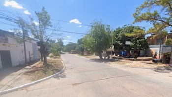 Vista de una calle residencial pavimentada, flanqueada por aceras, árboles y casas bajo un cielo azul con nubes dispersas