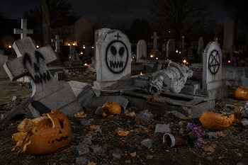Lápidas de piedra con grafitis negros, una cruz caída, una estatua rota y trozos de calabazas en el suelo de un cementerio de noche.