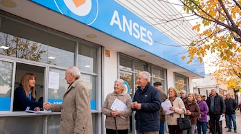 Una fila de adultos mayores espera para ser atendida en una oficina de ANSES. Una empleada entrega documentos a un jubilado en una ventanilla.