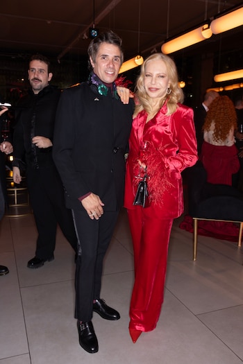 Un hombre y una mujer posan sonriendo. Él viste un traje oscuro con pañuelo de lentejuelas. Ella lleva un traje de terciopelo rojo con detalles de plumas
