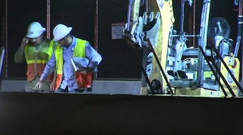Dos trabajadores con cascos blancos y chalecos de seguridad naranjas y verdes inspeccionan el suelo junto a una excavadora amarilla por la noche