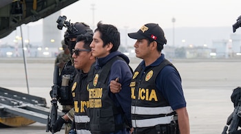 Un hombre detenido viste chaleco oscuro entre dos agentes de policía y un oficial armado cerca de un avión en una pista.