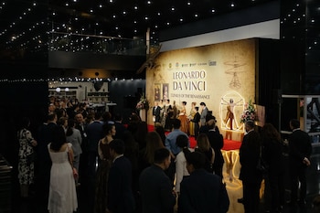 En un salón oscuro, una multitud observa un escenario iluminado con un cartel de la exposición 'Leonardo da Vinci' y varias personas en la tarima
