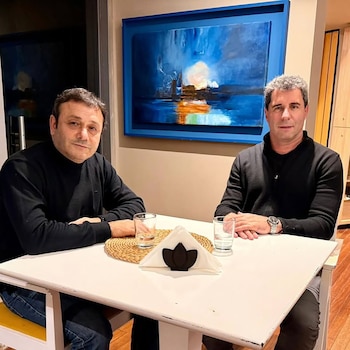 Dos hombres sentados frente a una mesa con dos vasos de agua, una servilleta, un posavasos tejido y un cuadro abstracto de tonos azules en la pared
