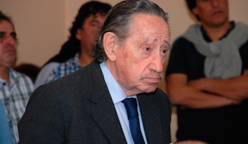 Primer plano de un hombre mayor con cabello largo canoso, bigote, traje oscuro, camisa celeste y corbata azul. Al fondo, personas desenfocadas