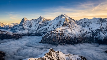 Cumbres de montañas rocosas y nevadas que emergen por encima de una capa de nubes bajo el cielo del amanecer.