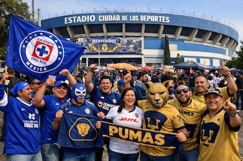 Multitud de aficionados de Cruz Azul y Pumas, algunos con banderas y máscaras, posan con entusiasmo frente al Estadio Ciudad de los Deportes bajo un cielo claro.