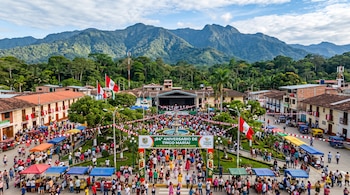 Vista elevada de una plaza decorada con banderas de Perú, puestos ambulantes, un arco publicitario y montañas en el fondo.