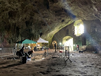 Interior de la Cueva Wogan en el Castillo de Pembroke, donde arqueólogos excavan el suelo. Se ven tiendas de campaña, equipo y luz entrando por ventanas arqueadas