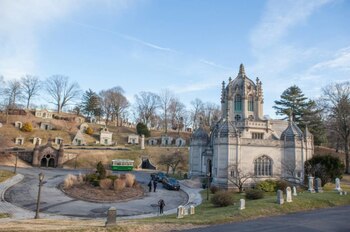 Vista de un cementerio con un gran edificio gótico de piedra en primer plano, flanqueado por mausoleos en una colina, caminos y árboles desnudos bajo un cielo azul