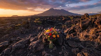 Un vibrante ramo de flores multicolores sobre rocas volcánicas oscuras al atardecer, con un volcán prominente bajo un cielo naranja y púrpura.