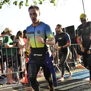 Un triatleta con traje de neopreno azul y amarillo mojado corre descalzo por un sendero al aire libre, con el público observando detrás de una valla