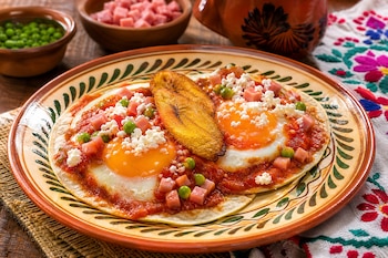 Dos huevos fritos sobre tortillas con salsa roja, jamón en cubos, guisantes, queso y plátano macho frito en un plato de cerámica decorado.