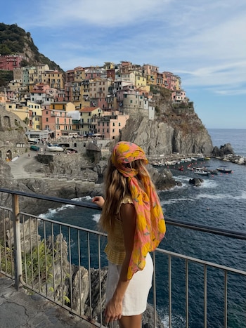 Mujer de espaldas con pañuelo en la cabeza, top amarillo y short blanco mira un pueblo costero con casas coloridas, un puerto, barcos y el mar