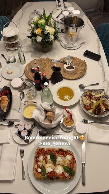 Una mesa de mármol blanco rectangular llena de platos de comida gourmet, incluyendo pizza, sopa, ensalada, postres, pan, champán en cubitera y un ramo de flores