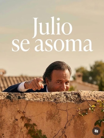 Julio Iglesias asomando la cabeza y un brazo por un muro de piedra, sonriendo y señalando con el dedo índice. El muro tiene plantas trepadoras