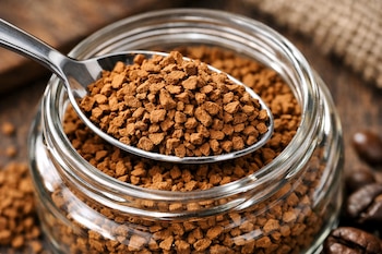 Una cuchara de metal con gránulos de café instantáneo, un frasco de vidrio lleno de café instantáneo y algunos granos de café enteros.