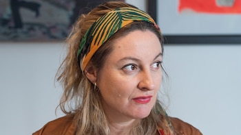 Primer plano de una mujer con una cinta verde y amarilla en el cabello, chaqueta marrón y lápiz labial rojo