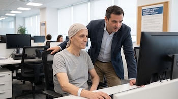 Dos hombres en una oficina; uno sentado con pañuelo en la cabeza y pulsera médica, el otro de pie a su lado mirando una pantalla de computadora.