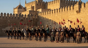 Soldados a caballo con lanzas y estandartes rojos forman una línea ordenada frente a un muro de fortaleza de piedra bajo la luz del atardecer.
