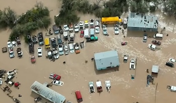Vista aérea de varias edificaciones y decenas de automóviles, furgonetas y un autobús rodeados por agua marrón en un terreno anegado