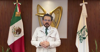 Zoé Robledo con anteojos y chaqueta frente a la bandera de México, la bandera del IMSS y un símbolo dorado sobre pared de madera