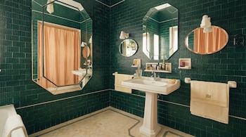 Vista de un baño con paredes de azulejos verde oscuro, un lavabo de pedestal blanco, espejos, toallas colgadas en toalleros y una cortina de ducha color melocotón.