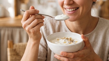 Mujer sonriendo come de un bol blanco con una cuchara de metal. El bol contiene una crema blanca con granola, semillas y miel