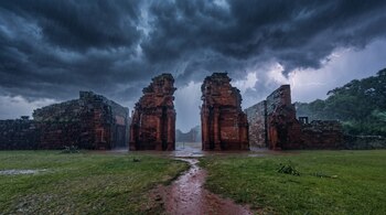 Ruinas de piedra roja bajo un cielo oscuro con lluvia, nubes densas y un relámpago en el fondo sobre césped mojado.