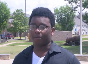 Un joven afroamericano con gafas y camisa negra, con un fondo de árboles verdes, edificios universitarios y un cielo soleado