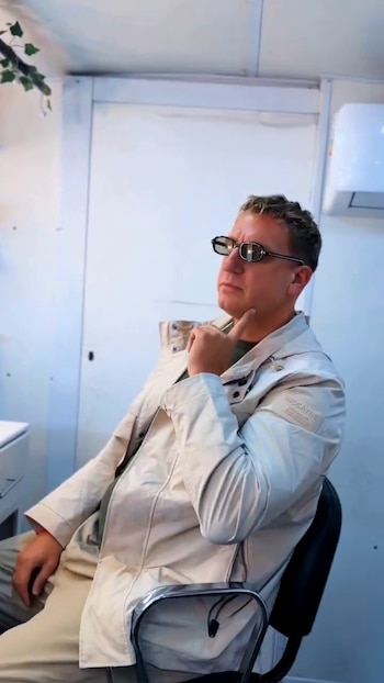 Un hombre con gafas de sol y chaqueta clara sostiene un dedo en la barbilla sentado en una silla negra