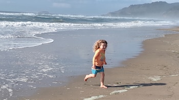 Un niño con camiseta naranja y pantalón corto azul corre por la arena de una playa con olas marinas y colinas al fondo.