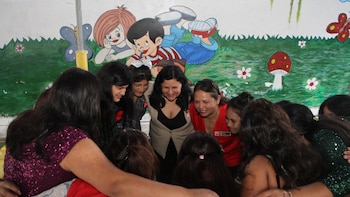 Doce mujeres y adolescentes sonrientes abrazándose en círculo frente a un mural infantil con dibujos de personas y naturaleza