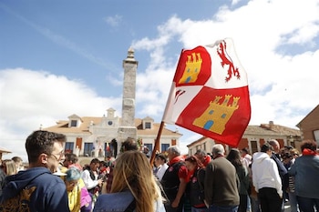 Vista de una multitud en una plaza con un monumento de piedra y edificios bajo un cielo azul. Una bandera roja y blanca con un león y castillos ondea en primer plano