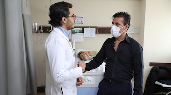 Un médico con bata blanca y un hombre con camisa negra y mascarilla blanca hacen un saludo de puño. Detrás, una mujer adulta en una cama de hospital.