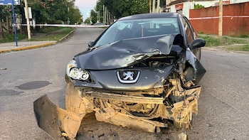 Coche Peugeot gris oscuro severamente dañado en la parte delantera, con capó arrugado y parachoques desprendido, en una calle con cinta policial