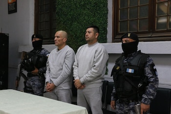 Dos hombres esposados y vestidos con ropa gris permanecen de pie junto a dos agentes de seguridad con el rostro cubierto