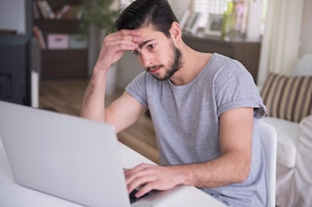Un hombre joven con barba viste una camiseta gris y toca su frente mientras mira la pantalla de una computadora portátil