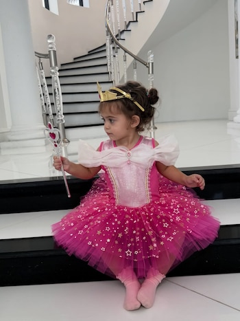 Niña de pelo castaño rizado con corona dorada y disfraz rosa de princesa, sentada en una escalera blanca y negra, sostiene una varita