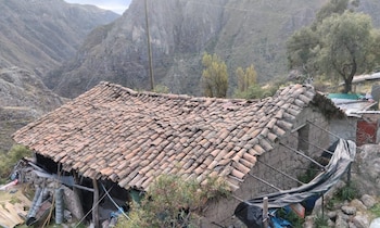 Casa rural con tejado de tejas de barro dañado y paredes de adobe agrietadas. Alrededor hay arbustos, árboles y montañas al fondo