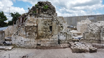 Estructuras de piedra en un sitio arqueológico con relieves esculpidos, vegetación superior, herramientas de trabajo y un cartel informativo.
